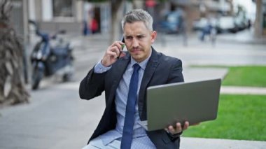 Genç İspanyol iş adamı sokakta dizüstü bilgisayar kullanarak akıllı telefondan konuşuyor.
