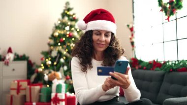 Orta yaşlı İspanyol kadın akıllı telefon ve kredi kartı kullanıyor. Evdeki Noel ağacının yanında koltukta oturuyor.