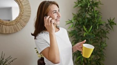 Elinde bir fincan kahve tutan genç bir kadın evde akıllı telefondan konuşuyor.