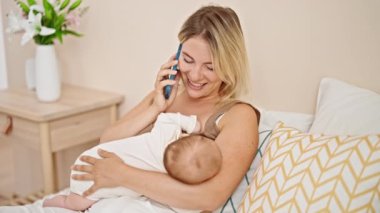 Anne ve kızı yatakta oturup bebeği emziriyorlar. Yatak odasında akıllı telefondan konuşuyorlar.