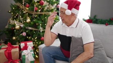 Orta yaşlı, gri saçlı bir adam Noel ağacının yanındaki koltukta oturmuş evde sıkılmış görünüyor.