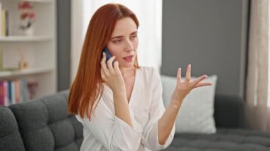 Genç kızıl saçlı kadın evde ciddi bir ifadeyle akıllı telefondan konuşuyor.