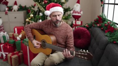 Genç kel adam evdeki noel ağacının yanındaki koltukta klasik gitar çalıyor.