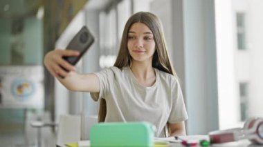 Genç güzel kız öğrenci kütüphanede gülümseyen akıllı telefonla selfie çekiyor.