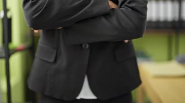 Genç, güzel, İspanyol bir iş kadını. Kollarını kavuşturup ofiste duruyor.