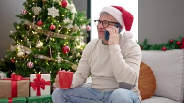 Orta yaşlı, kır saçlı, akıllı telefondan konuşan, Noel ağacının yanında oturup kahve içen bir adam.