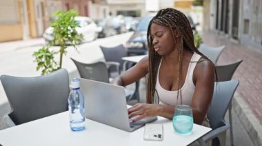 Afro-Amerikalı bir kadın kahve dükkanının terasında bilgisayarla akıllı telefondan konuşuyor.