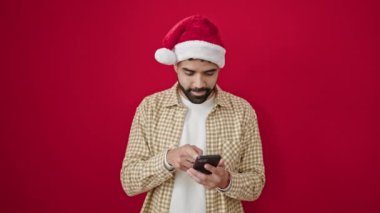Akıllı telefon ve kredi kartıyla alışveriş yapan genç İspanyol adam izole edilmiş kırmızı arka planda noel şapkası takıyor.