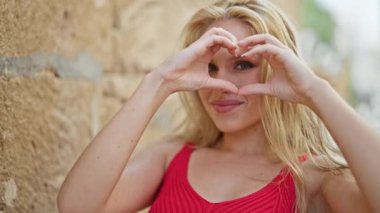 Young blonde woman smiling confident doing heart gesture with hands at street