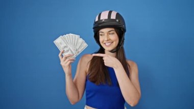 Motosiklet kaskı takan genç ve güzel bir İspanyol kadın dolarları işaret ederek, izole edilmiş mavi arka planda baş parmak hareketi yapıyor.
