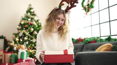 Sarışın bir kadın hediye paketini açıyor evdeki Noel ağacının yanındaki koltukta oturuyor.