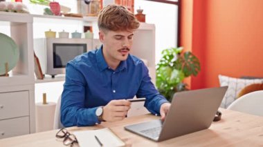 Laptop ve kredi kartıyla alışveriş yapan İspanyol genç yemek odasında üzgün görünüyor.