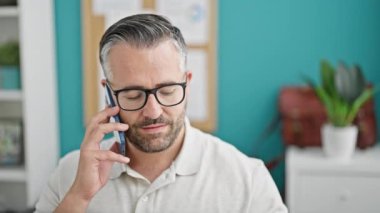 Gri saçlı iş adamı. Akıllı telefondan konuşuyor. Ofise bakıyor.