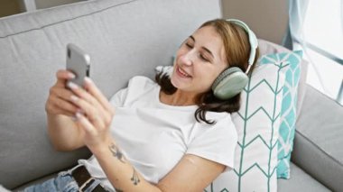 Akıllı telefon kullanan, kulaklık takan, evdeki kanepede yatan genç bir kadın.