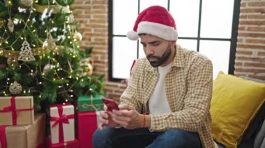 Genç İspanyol adam Noel şapkası takıyor. Akıllı telefon kullanıyor. Evde stresli.