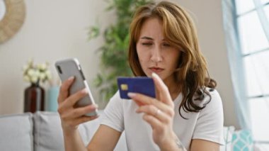 Akıllı telefon ve kredi kartıyla alışveriş yapan genç bir kadın evdeki kanepede oturuyor.