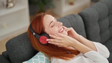 Genç kızıl saçlı kadın evdeki kanepede oturmuş müzik dinliyor.