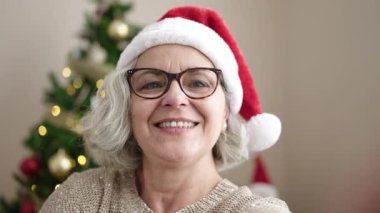 Gri saçlı orta yaşlı bir kadın Noel ağacının yanında video çağrısı yapıyor.