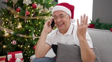 Orta yaşlı, kır saçlı bir adam evde Noel ağacının yanında telefonla konuşuyor.
