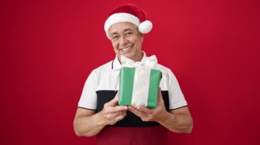 Orta yaşlı, gri saçlı, sırıtan, kendine güvenen, izole kırmızı arka planda bir Noel şapkası takan bir adam.