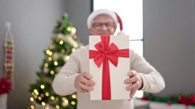 Orta yaşlı, kır saçlı bir adam elinde hediyesiyle Noel ağacının yanında duruyor.