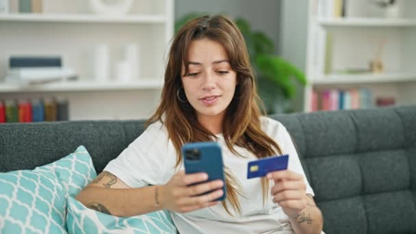 若い美しいヒスパニック系女性が自宅でソファーに座っているクレジットカードを持っているスマートフォンでセルフィーを作ります — ストック動画