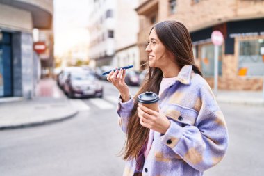 Young beautiful hispanic woman talking on smartphone drinking coffee at street