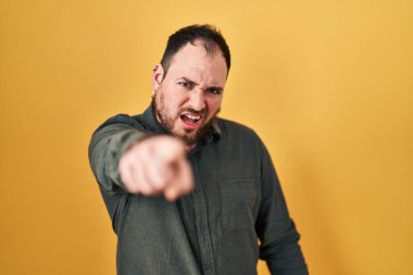 Plus size hispanic man with beard standing over yellow background pointing displeased and frustrated to the camera, angry and furious with you 