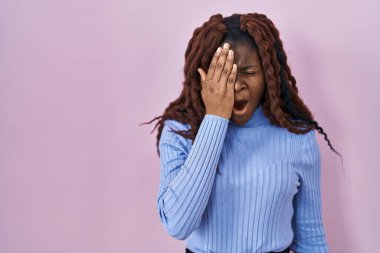 Pembe arka planda duran Afrikalı kadın. Yarı yüz, göz ve ağız kısmını elleriyle kapatmaktan yorulmuş. Yüzüm acıyor.. 