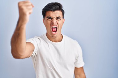Hispanic man standing over blue background angry and mad raising fist frustrated and furious while shouting with anger. rage and aggressive concept. 