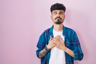 Pembe arka planda sakallı, elleri göğsünde, gözleri kapalı ve yüzünde minnet dolu bir jestle gülümseyen genç bir İspanyol. sağlık kavramı. 
