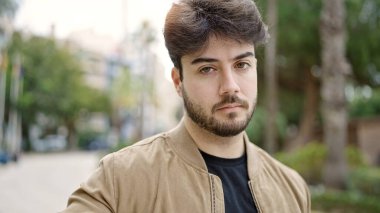 Young hispanic man standing with serious expression at park