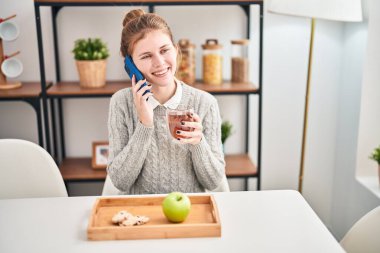 Sağlıklı kahvaltı yapan genç sarışın kadın yemek odasında akıllı telefondan konuşuyor.