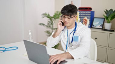Genç İspanyol doktor laptopunu kullanıyor. Klinikte akıllı telefondan konuşuyor.