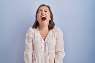 Middle age hispanic woman standing over blue background angry and mad screaming frustrated and furious, shouting with anger. rage and aggressive concept. 