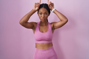 Pembe arka planda spor kıyafetleri giyen Afrikalı Amerikalı bir kadın boynuzlarını başlarının üzerinde gezdirirken komik hareketler yapıyor. 