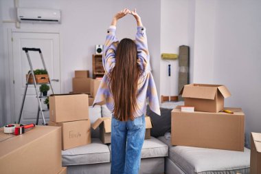 Young beautiful hispanic woman stretching arms standing on back view at new home