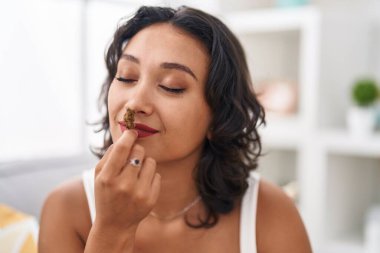 Young beautiful hispanic woman smelling marihuana plant sitting on sofa at home