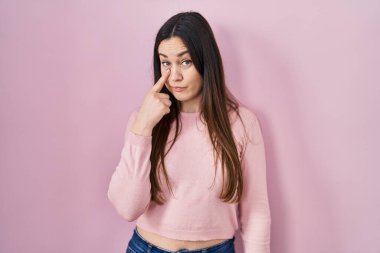 Pembe arka planda duran genç esmer kadın jestini izlerken gözüne işaret ediyor. Şüpheli bir ifade. 