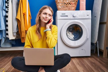 Young blonde woman using laptop talking on smartphone waiting for washing machine at laundry room