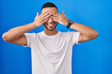 Mavi arka planda dikilen genç İspanyol adam, neşeli ve komik gülümseyen ellerle gözlerini kapatıyor. kör kavram. 