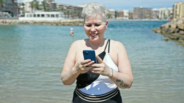 Kumsalda akıllı telefon kullanarak mayo giyen orta yaşlı, kır saçlı bir kadın turist.
