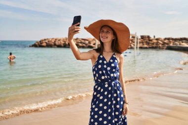 Yaz şapkası takan genç sarışın kadın sahilde akıllı telefondan selfie çekiyor.