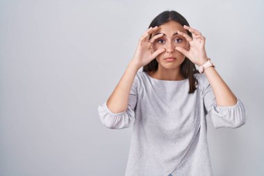 Young hispanic woman standing over white background trying to open eyes with fingers, sleepy and tired for morning fatigue 