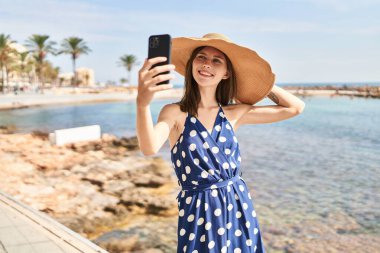 Yaz şapkası takan genç sarışın kadın sahilde akıllı telefondan selfie çekiyor.