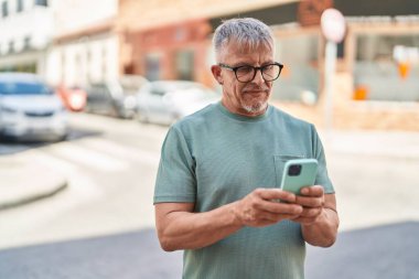 Orta yaşlı, kır saçlı, kendine güvenen, caddede akıllı telefon kullanan bir adam.