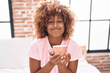 African american woman smelling cup of coffee sitting on bed at bedroom