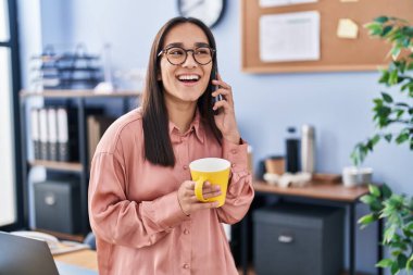 Young hispanic woman business worker talking on smartphone drinking coffee at office