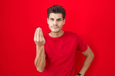 Genç İspanyol adam kırmızı arka planda duruyor. İtalyan jesti yapıyor. El ve parmakları kendinden emin bir ifadeyle. 