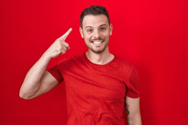 Young hispanic man standing over red background smiling pointing to head with one finger, great idea or thought, good memory 